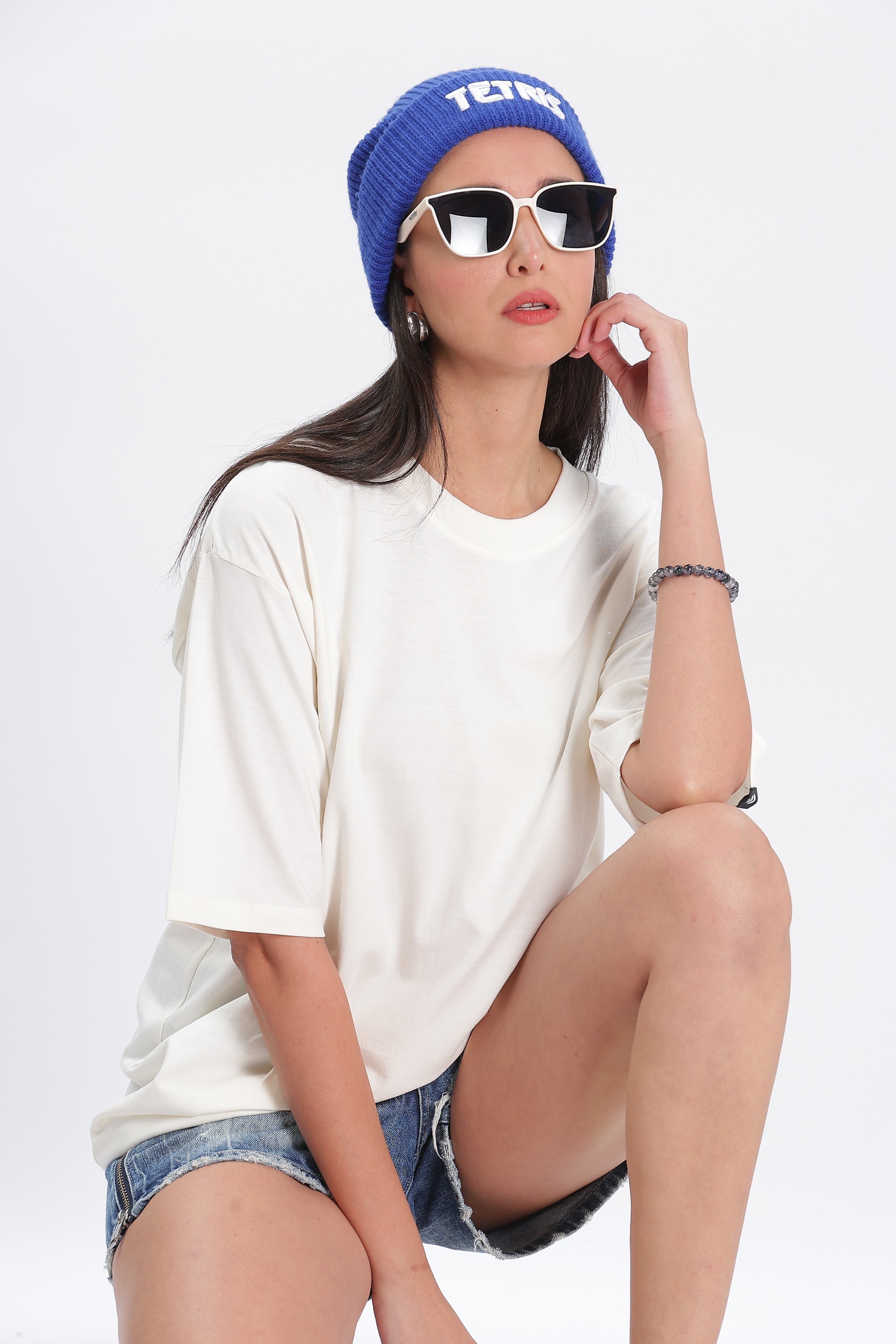 Woman wearing oversized solid t shirts for women in off-white with sunglasses and blue beanie sitting confidently
