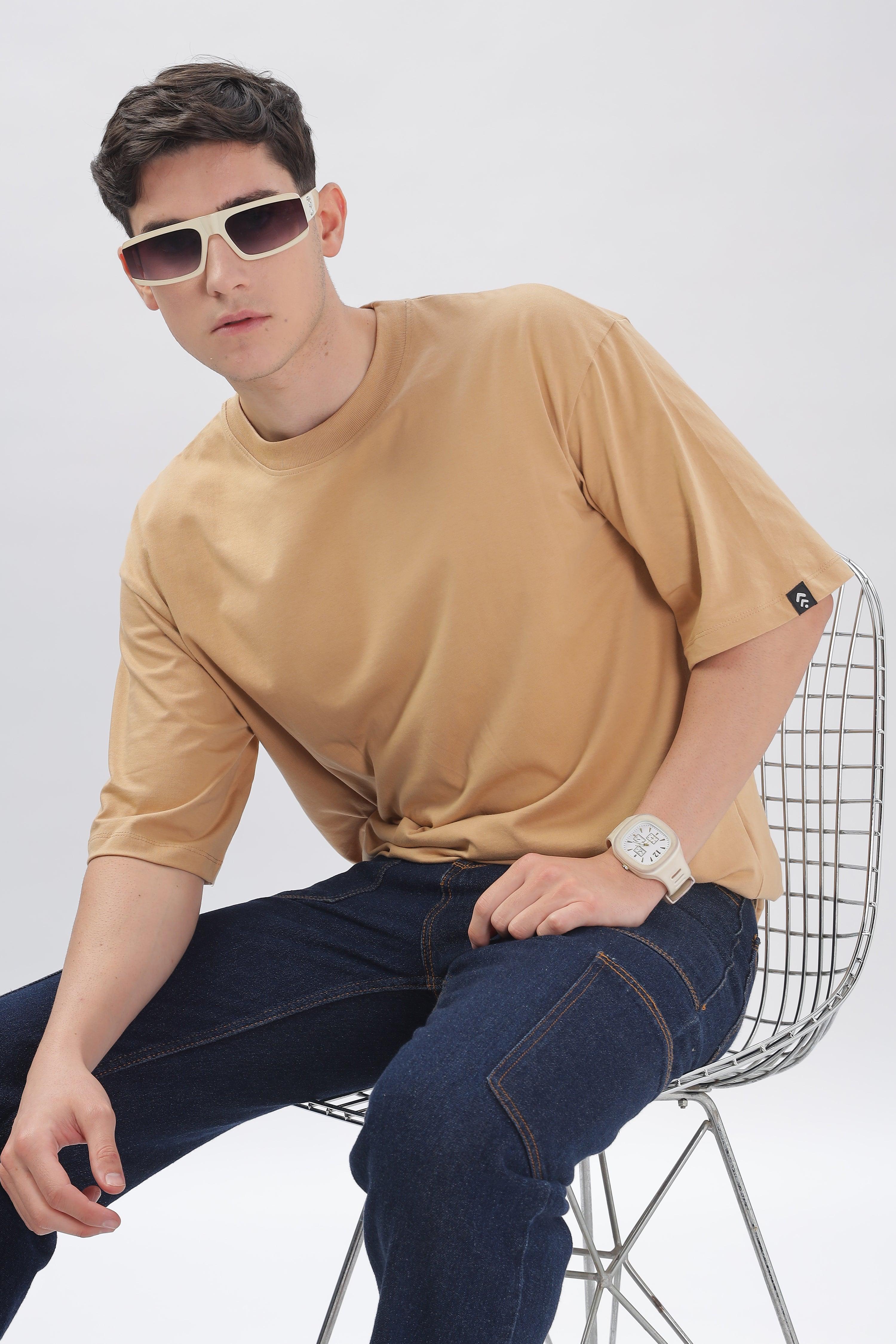 Male model wearing taupe oversized t-shirt by Soulmonk seated on a wire chair