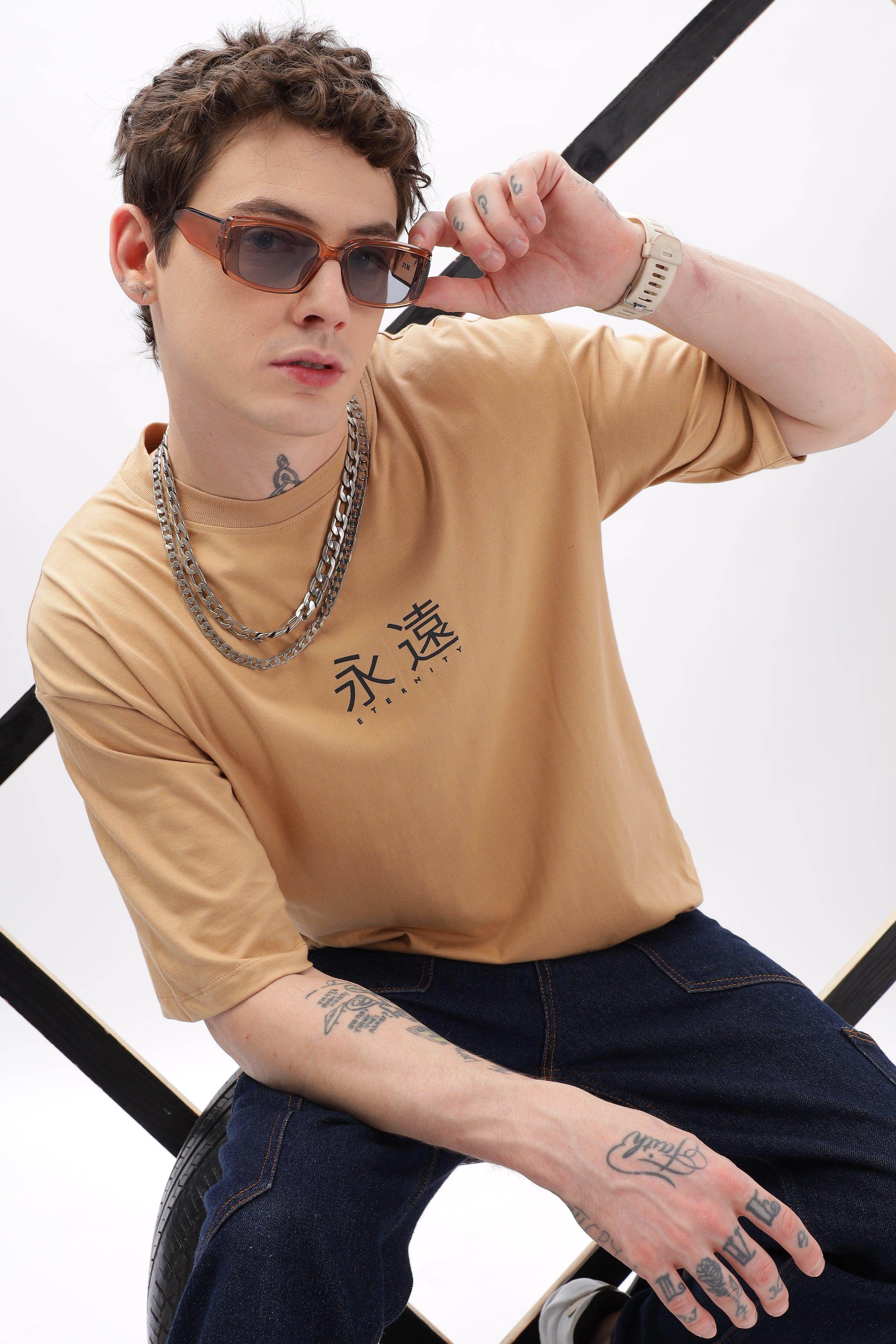 Male model wearing a taupe oversized t-shirt with black graphic text paired with dark jeans and sunglasses
